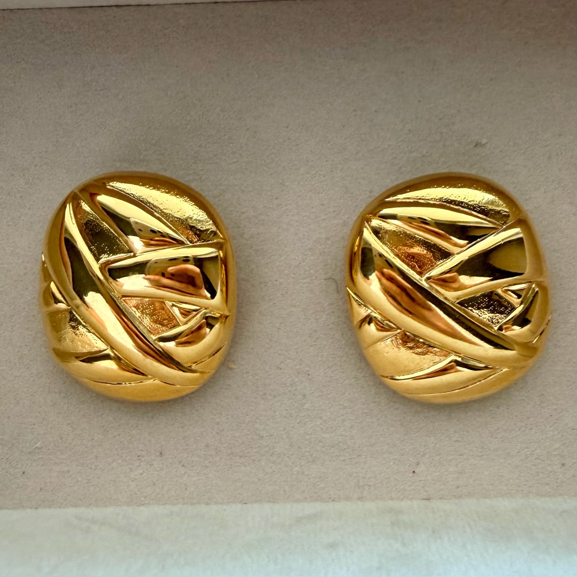A pair of round gold earrings with an abstract, textured pattern, displayed on a light beige textured surface.