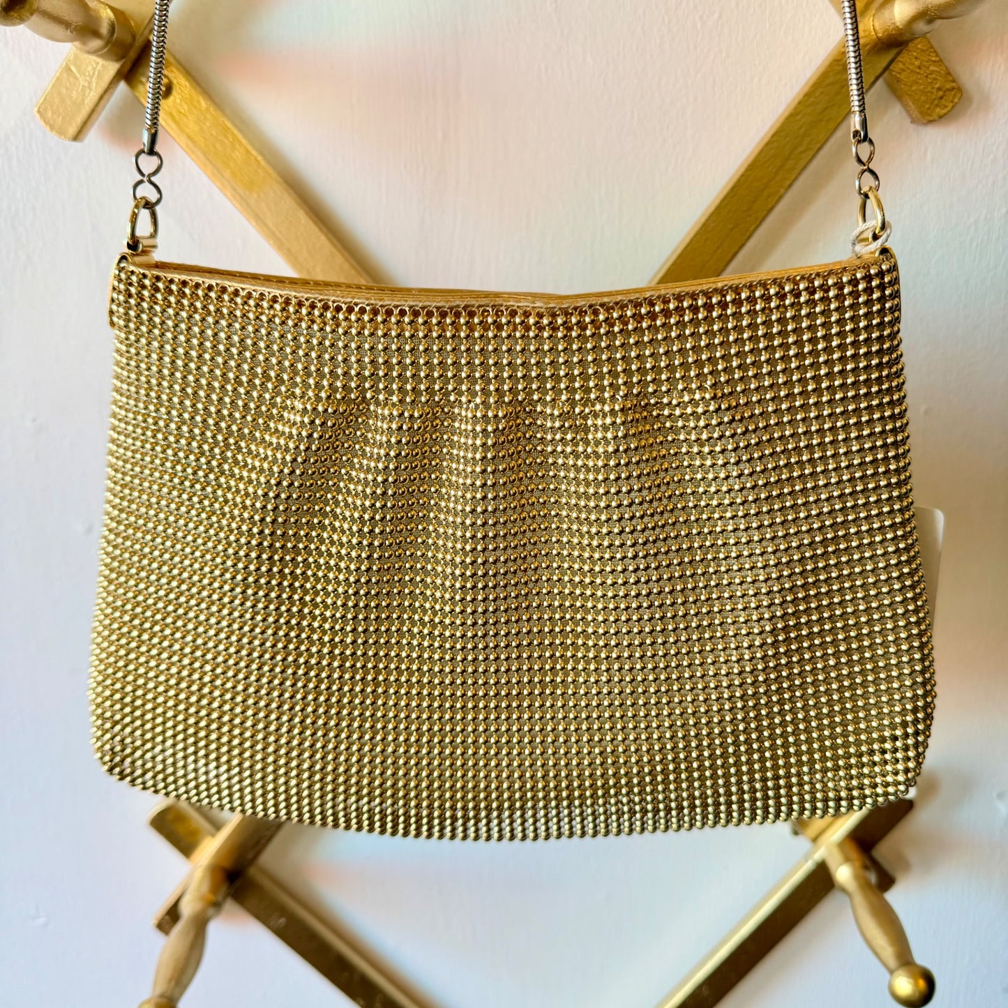 A gold-tone mesh chain purse with a silver-tone chain strap, hanging from a gold metal stand against a white wall.
