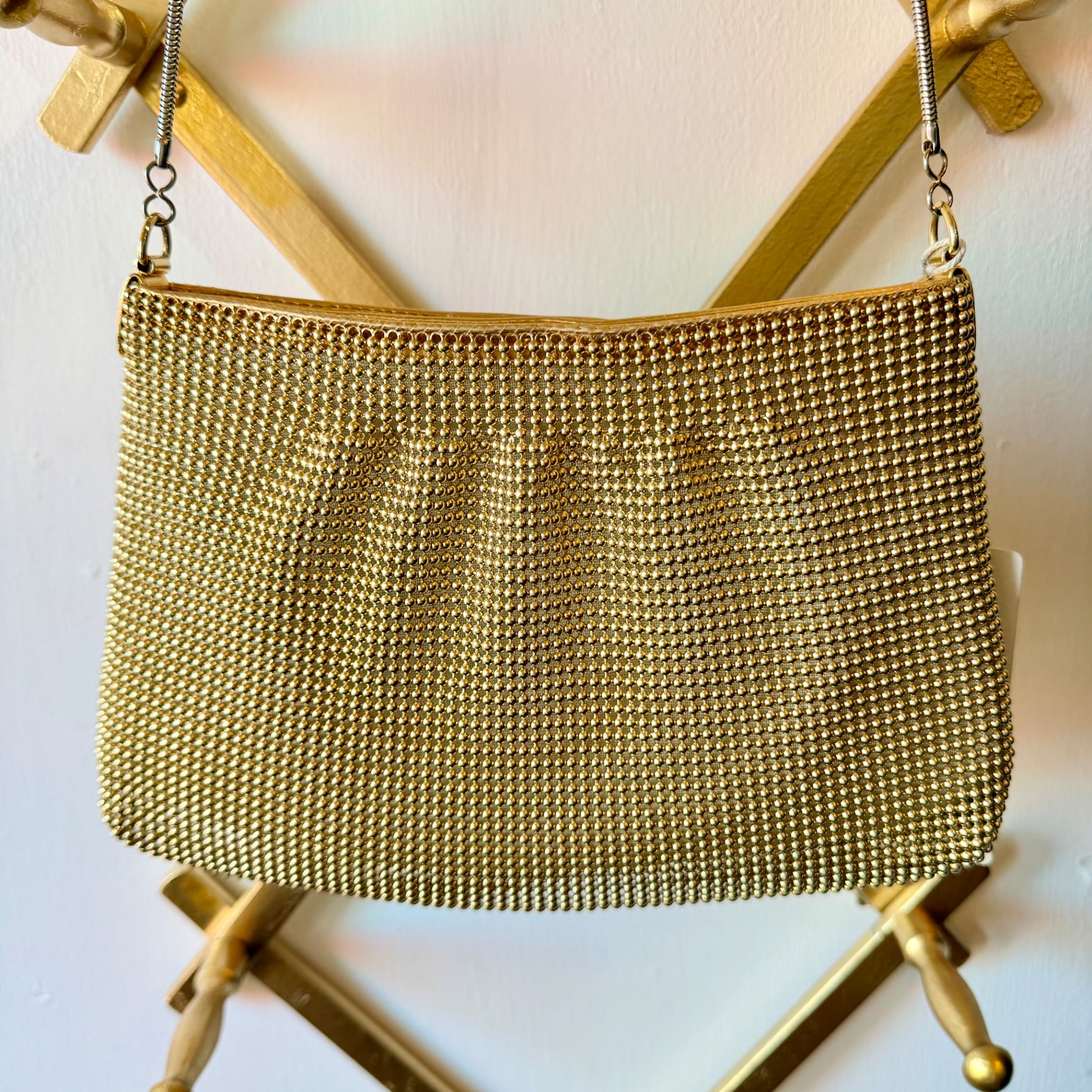 A gold-tone mesh chain purse with a silver-tone chain strap, hanging from a gold metal stand against a white wall.