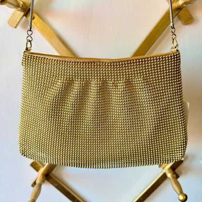 A gold-tone mesh chain purse with a silver-tone chain strap, hanging from a gold metal stand against a white wall.