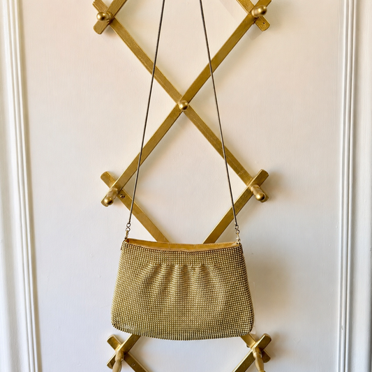 A gold mesh purse with a thin shoulder strap hanging from a gold wall-mounted geometric rack against a white paneled wall.