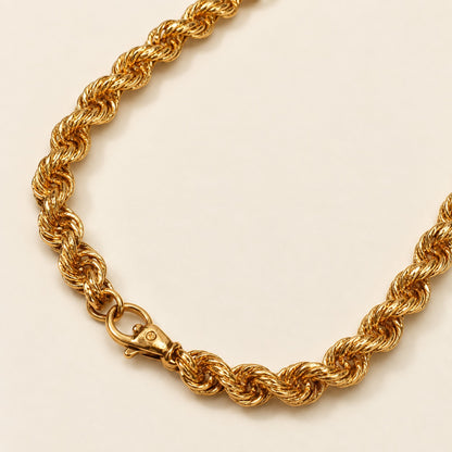 A close-up of a gold rope chain necklace with a lobster clasp, displayed on a clean, solid cream background.