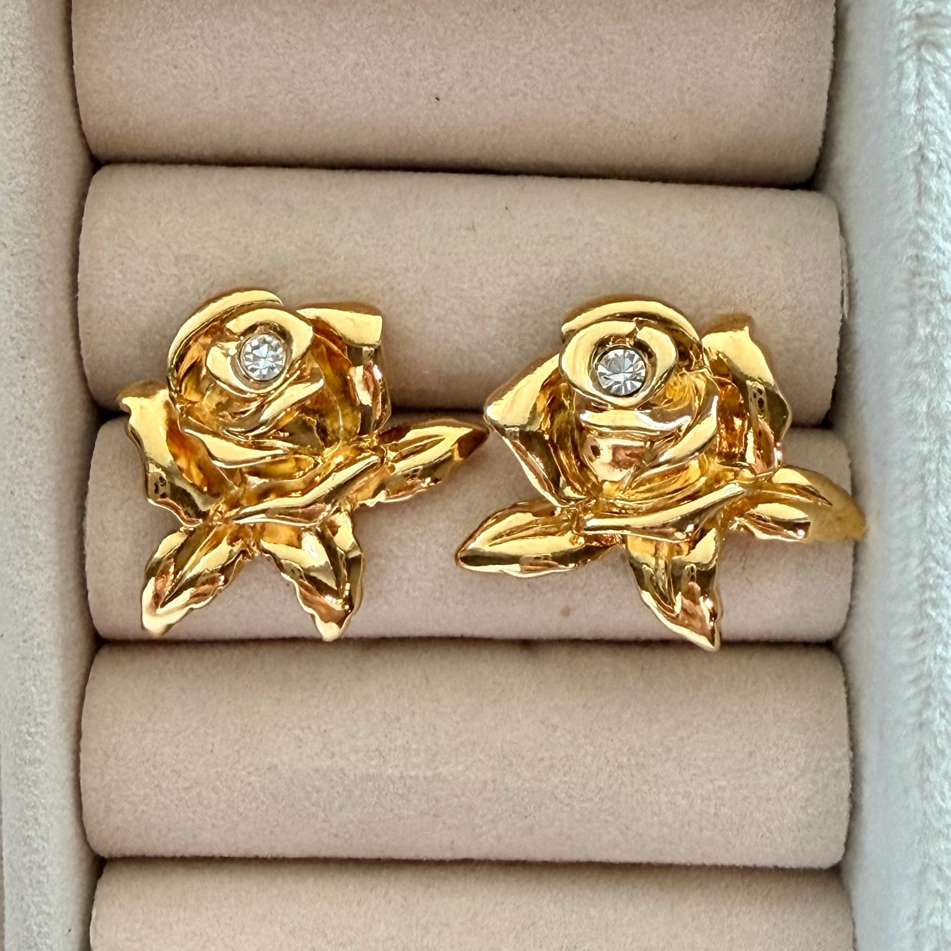A pair of gold rose-shaped stud earrings with small gemstones in the center, displayed on a beige jewelry box cushion.