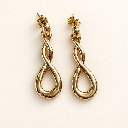 A pair of gold drop earrings with an infinity symbol design, photographed on a clean, solid off-white background.