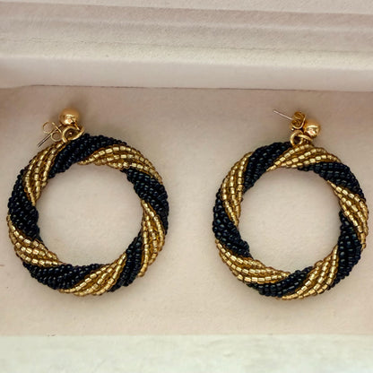 A pair of handmade beaded hoop earrings with a twisted spiral pattern in black and gold, displayed inside a white jewelry box.