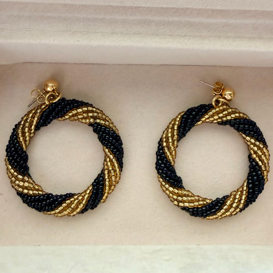 A pair of handmade beaded hoop earrings with a twisted spiral pattern in black and gold, displayed inside a white jewelry box.