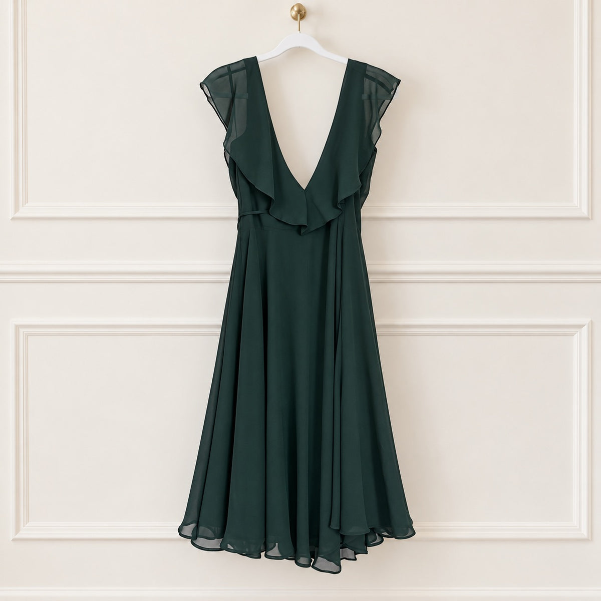 A dark forest green sleeveless chiffon dress with a deep V-neckline, ruffled yoke, and a flared skirt, hanging on a white hanger against a cream-colored paneled wall.