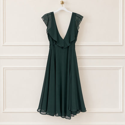 A dark forest green sleeveless chiffon dress with a deep V-neckline, ruffled yoke, and a flared skirt, hanging on a white hanger against a cream-colored paneled wall.