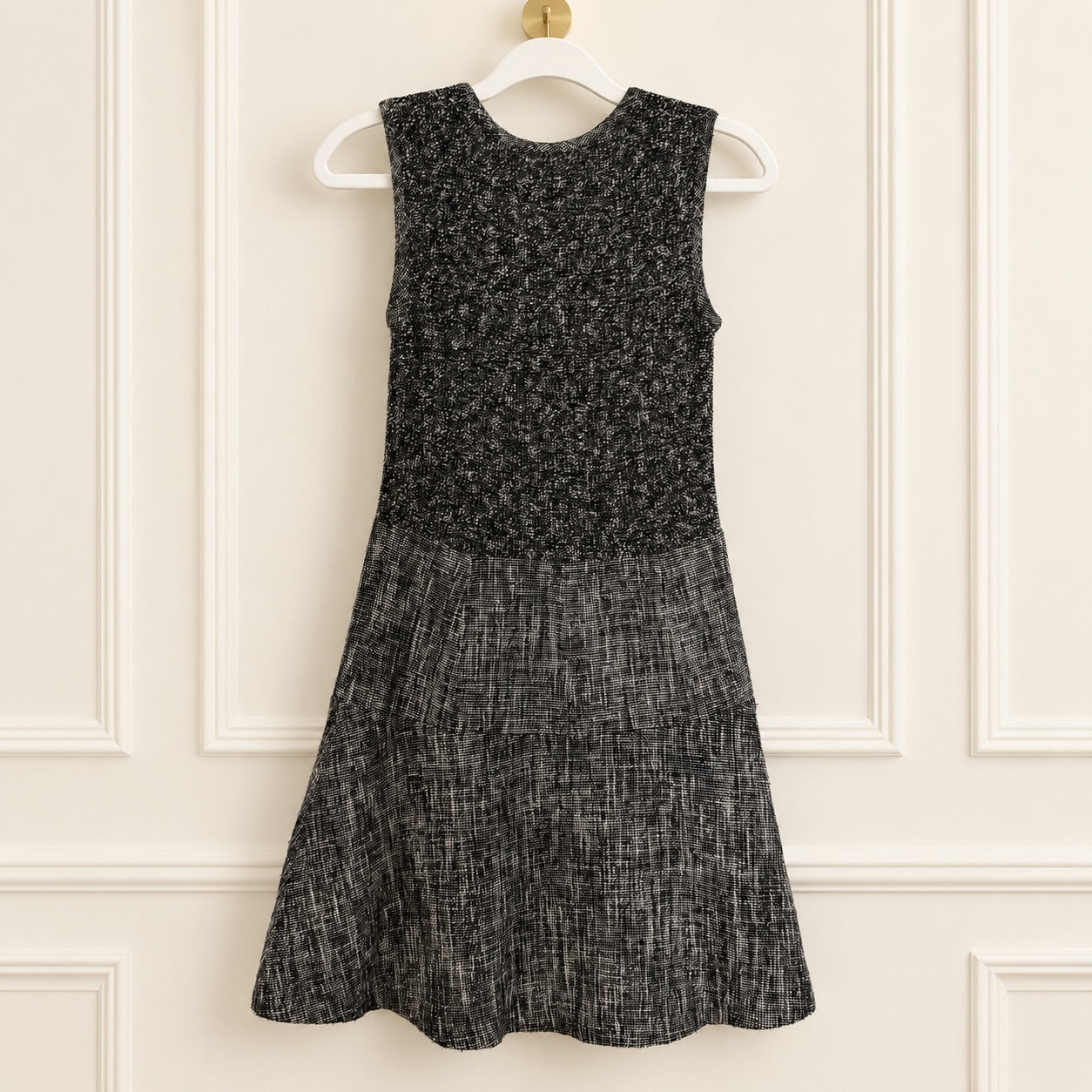 A sleeveless black and gray textured dress hanging on a white hanger against a cream paneled wall.