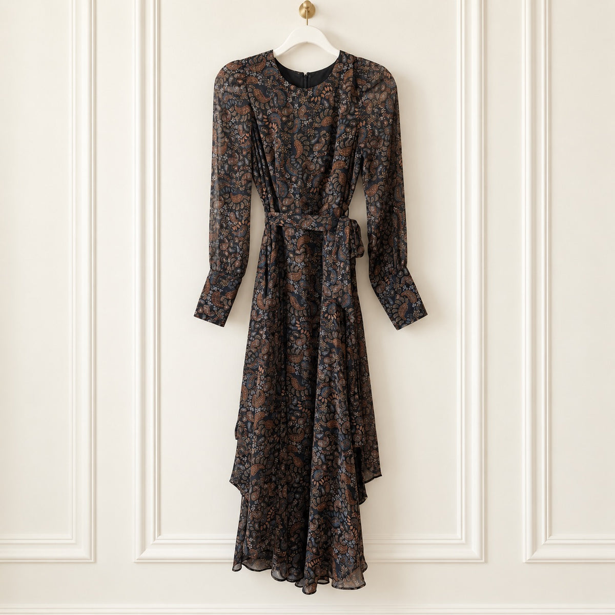 A long-sleeve midi dress with a dark brown and black floral pattern, featuring a tie waist belt, hanging on a white hanger against a white paneled wall.