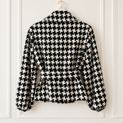 A black and white houndstooth patterned long-sleeve jacket hanging on a gold hanger against a white paneled wall.