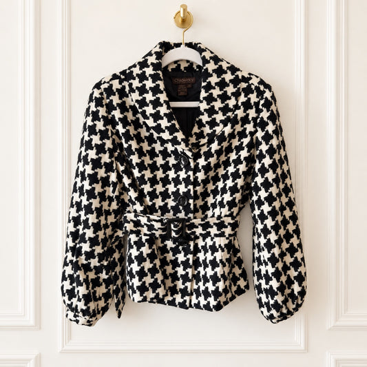 A black and cream houndstooth belted jacket hanging on a white hanger with a gold ring against a white paneled wall.
