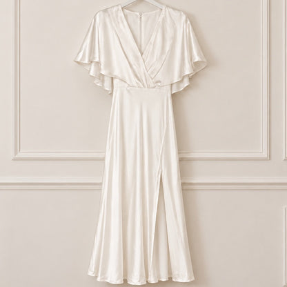 An ivory satin wrap dress with flutter sleeves hangs on a white hanger against a cream-colored paneled wall.