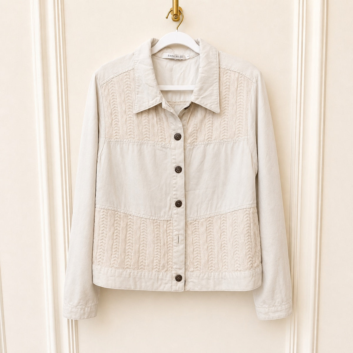 A light beige cropped jacket with textured cable-knit panels on the chest and lower front, hanging on a white hanger against a cream paneled door.