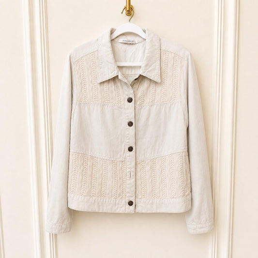 A light beige cropped jacket with textured cable-knit panels on the chest and lower front, hanging on a white hanger against a cream paneled door.