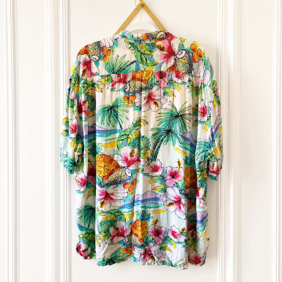 A short-sleeve button-up shirt with a vibrant tropical Hawaiian print, hanging on a wooden hanger against a white paneled door.