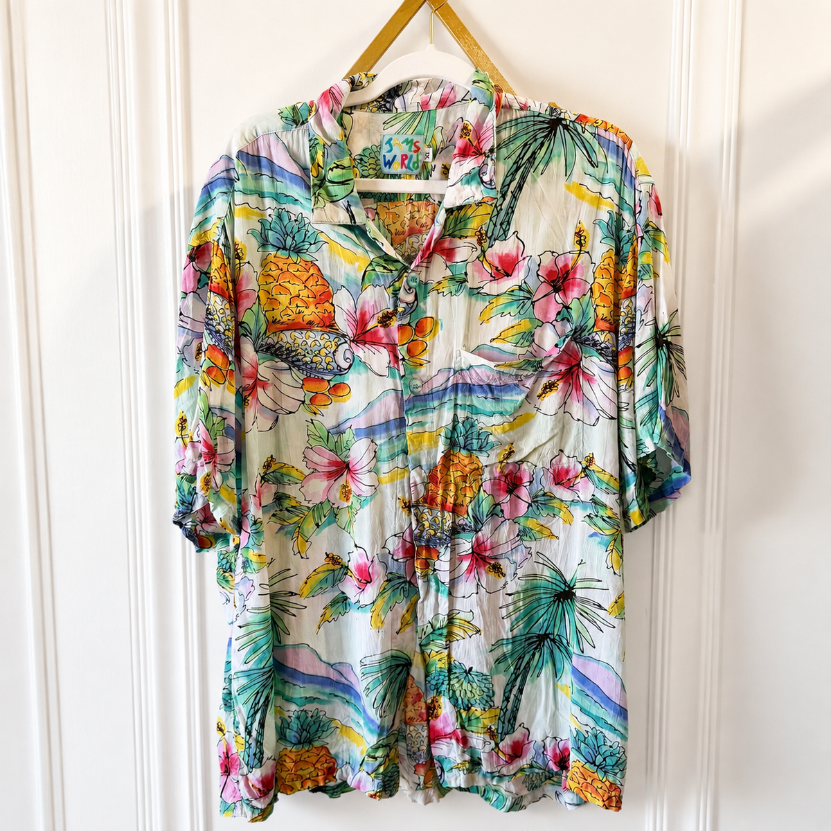 A short-sleeve button-up shirt with a vibrant tropical print, hanging on a wooden hanger against a white paneled door. The print features pink hibiscus flowers, green palm leaves, pineapples, and colorful scenic motifs on a cream-colored base.