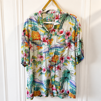 A short-sleeve button-up shirt with a vibrant tropical print, hanging on a wooden hanger against a white paneled door. The print features pink hibiscus flowers, green palm leaves, pineapples, and colorful scenic motifs on a cream-colored base.