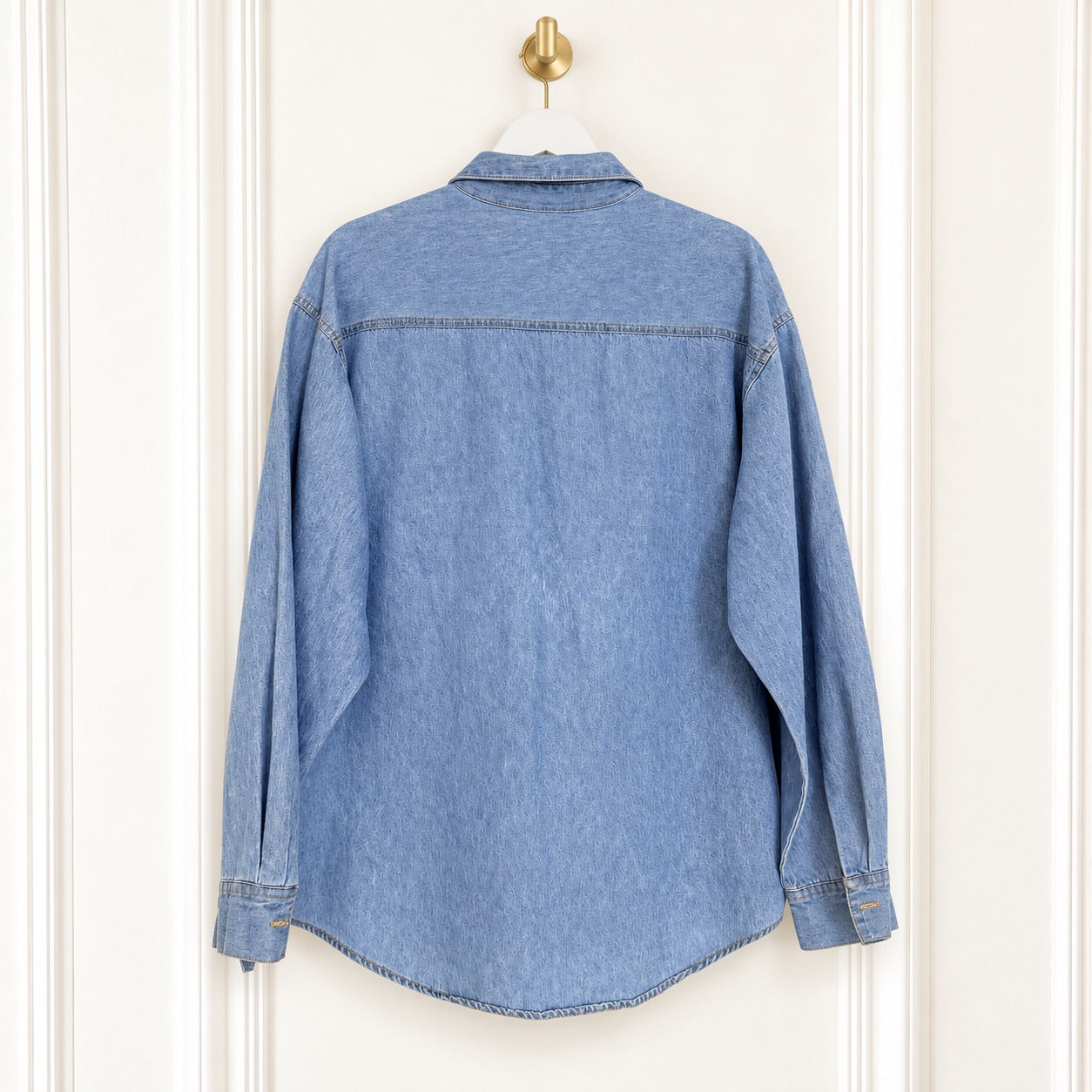A light blue denim shirt jacket hanging on a white hanger against a white paneled door, showing the back view.