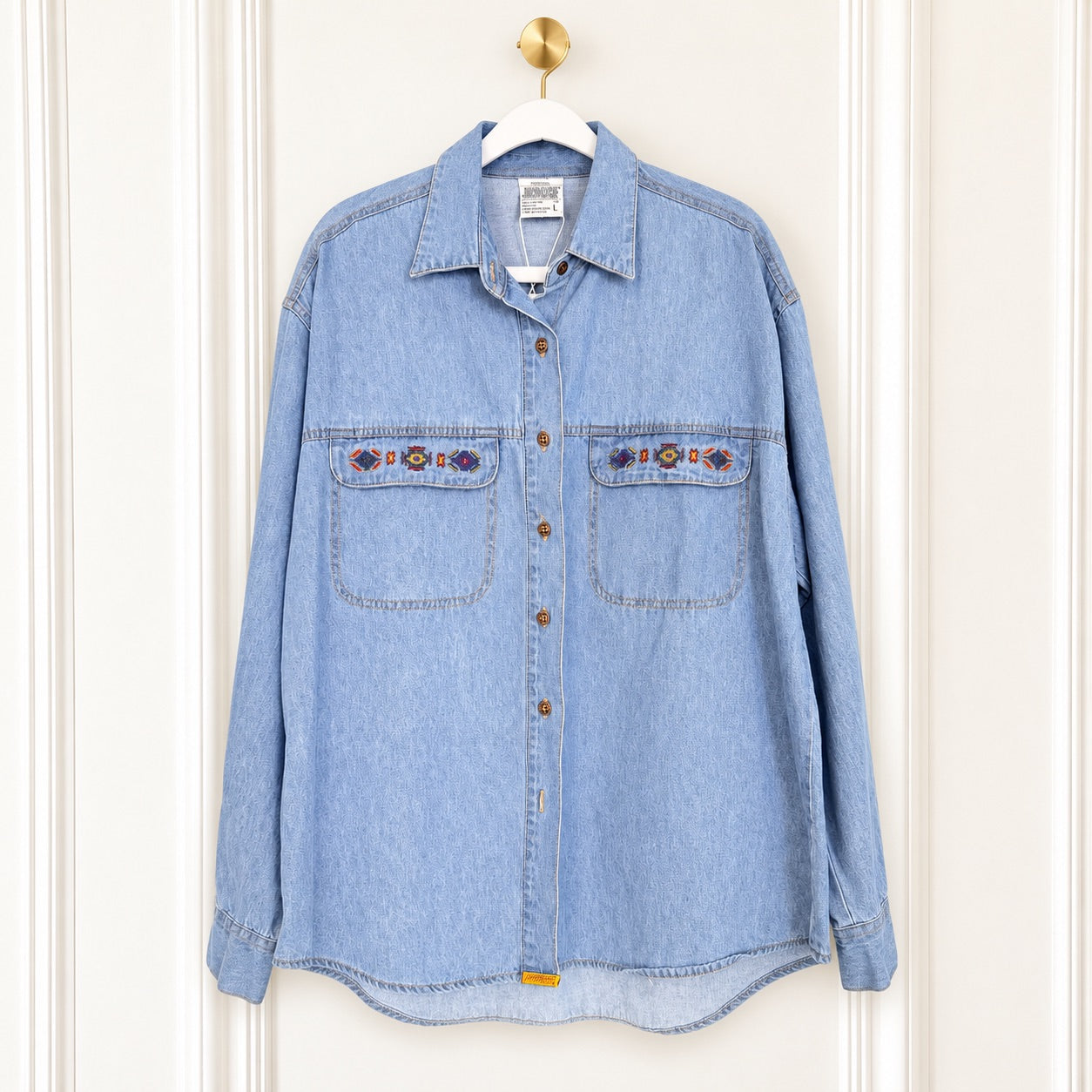 A light blue denim button-up shirt with two chest pockets and embroidered details, hanging on a white hanger against a white paneled door.