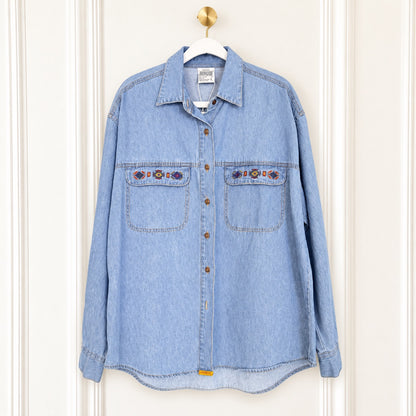 A light blue denim button-up shirt with two chest pockets and embroidered details, hanging on a white hanger against a white paneled door.