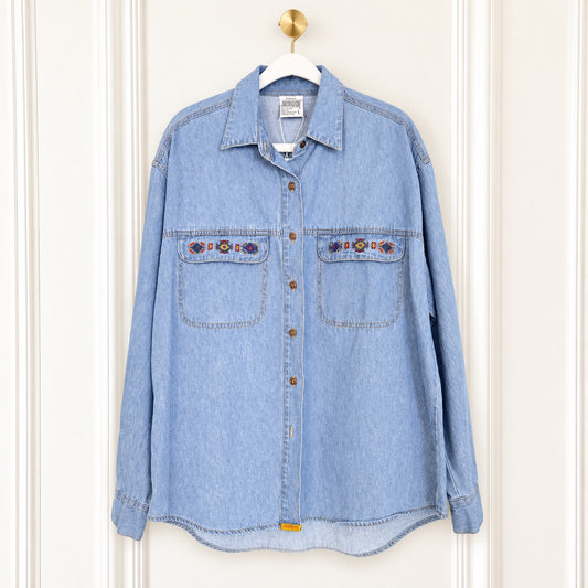 A light blue denim button-up shirt with two chest pockets and embroidered details, hanging on a white hanger against a white paneled door.