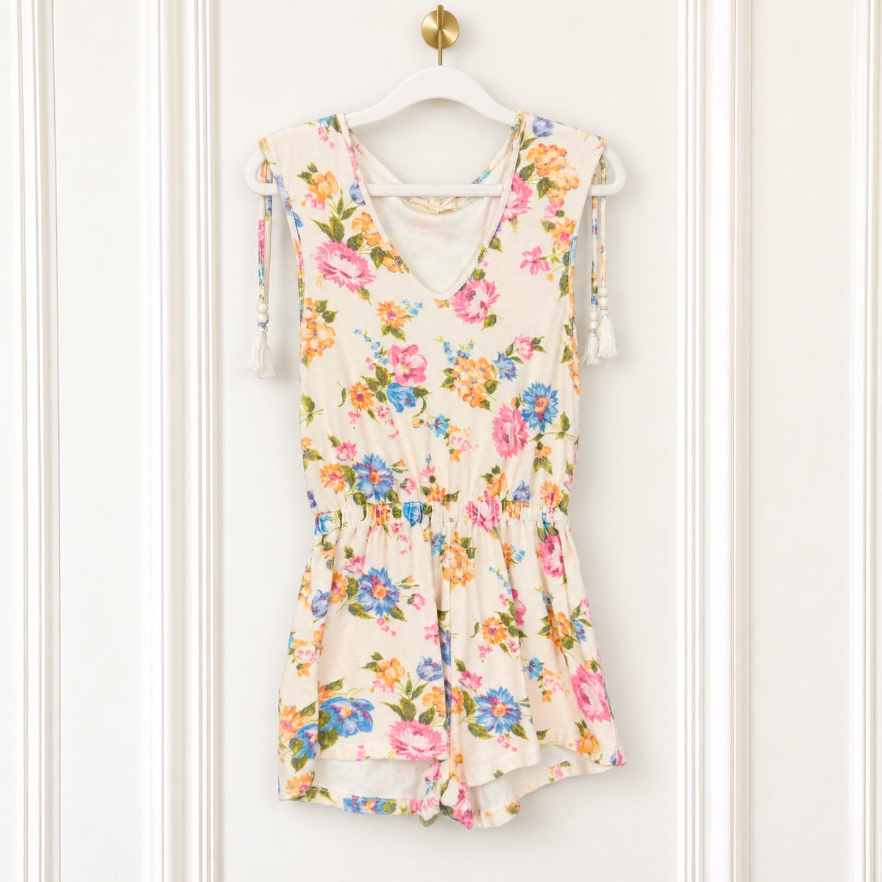 A sleeveless cream-colored romper with a vibrant pink, blue, and orange floral print, hanging on a white hanger against a white paneled door.