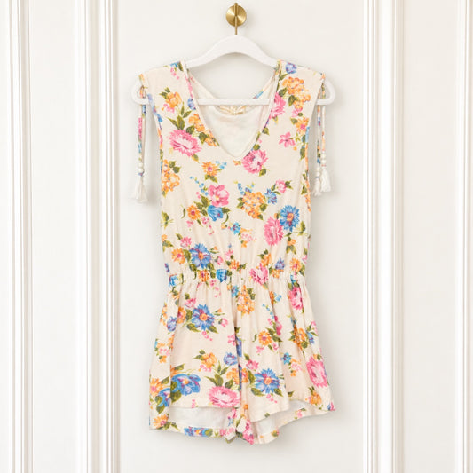 A sleeveless cream-colored romper with a vibrant pink, blue, and orange floral print, hanging on a white hanger against a white paneled door.