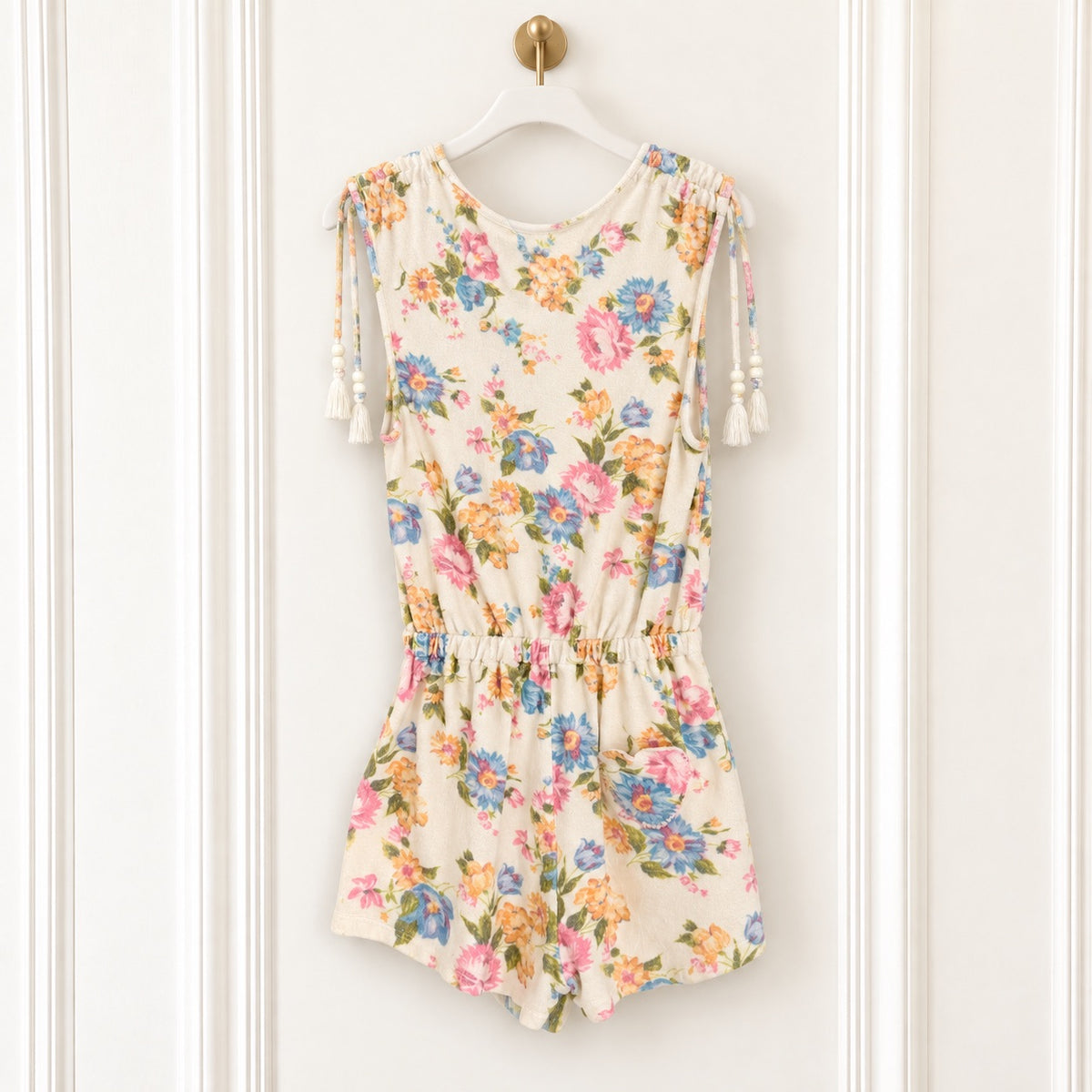 A sleeveless cream-colored romper with a colorful floral print, featuring tassel ties on the shoulders and a gathered elastic waist, hanging on a white hanger against a white paneled wall.