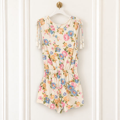 A sleeveless cream-colored romper with a colorful floral print, featuring tassel ties on the shoulders and a gathered elastic waist, hanging on a white hanger against a white paneled wall.