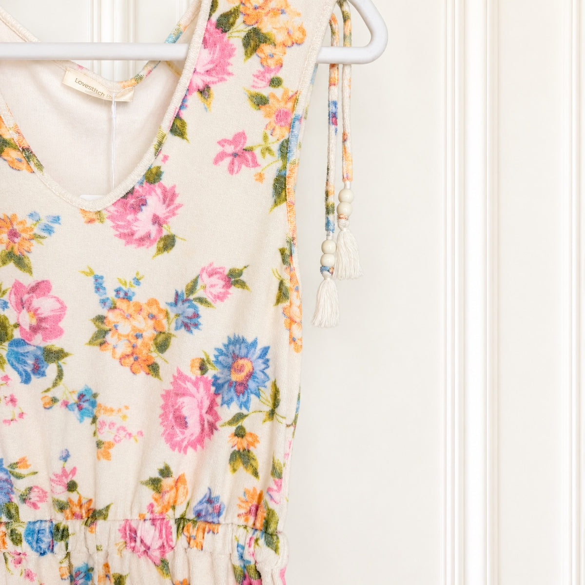 A sleeveless cream-colored dress with a colorful floral print, hanging on a white hanger against a white paneled door. The dress features tie-shoulder straps with tassels and a gathered waist.