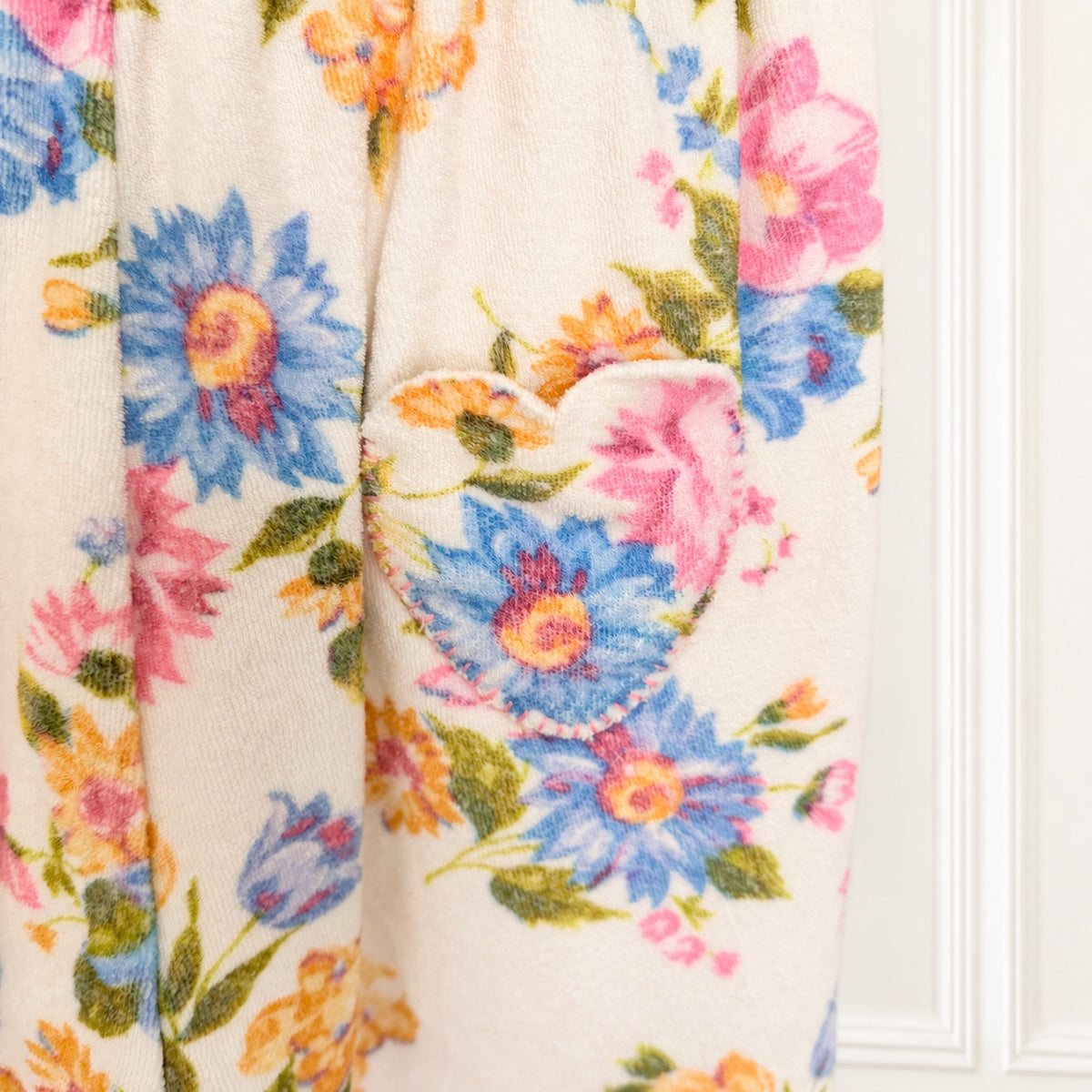 A close-up of a cream-colored fleece blanket featuring a colorful floral pattern with blue, pink, and orange flowers, and a heart-shaped patch pocket.