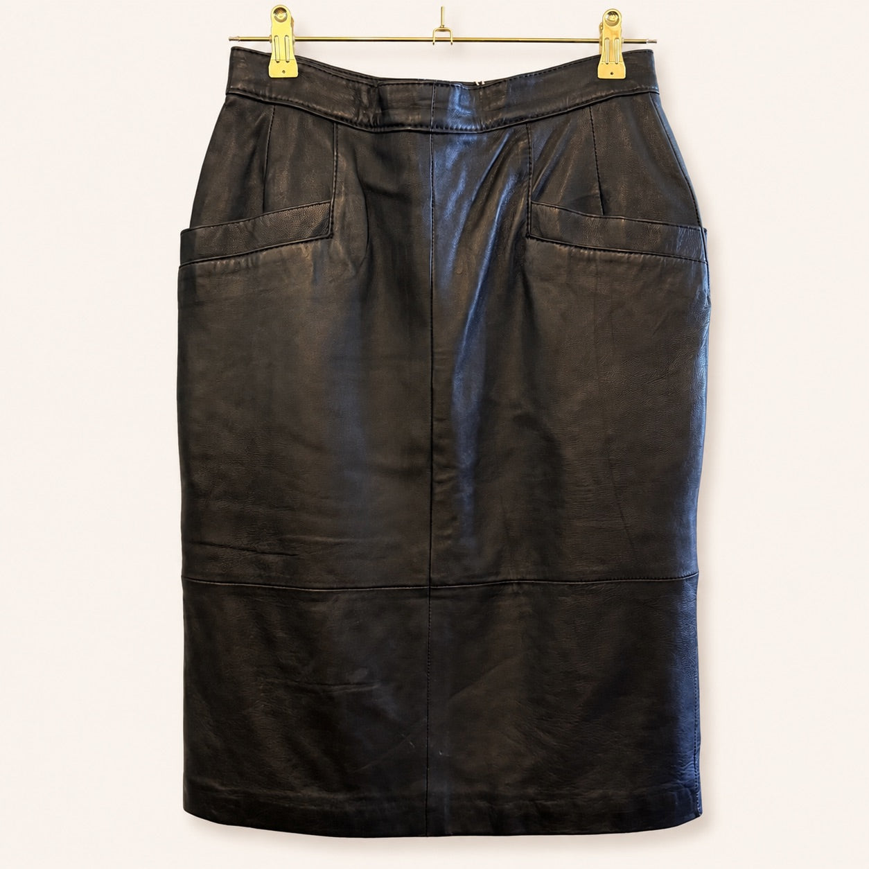 A black leather pencil skirt with two large front patch pockets, hanging from a gold metal hanger with gold clips against a plain off-white background.