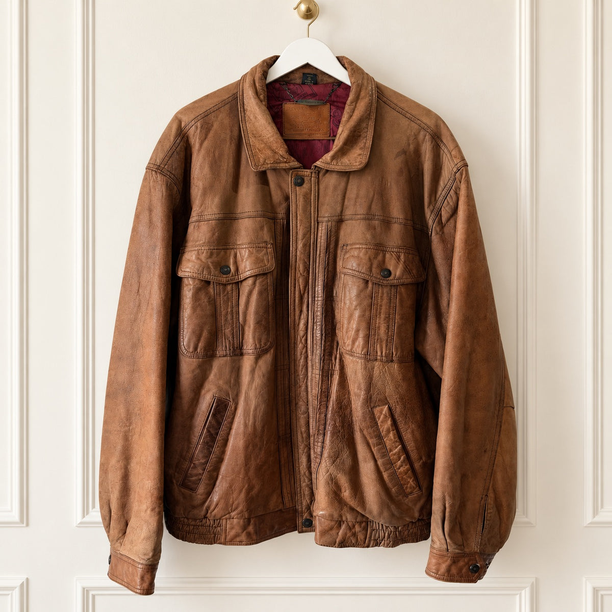 A brown leather jacket with a distressed finish, featuring a front zipper, two large chest flap pockets, and two lower hand-warmer pockets, hanging on a white hanger against a white paneled wall.
