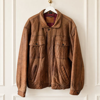 A brown leather jacket with a distressed finish, featuring a front zipper, two large chest flap pockets, and two lower hand-warmer pockets, hanging on a white hanger against a white paneled wall.