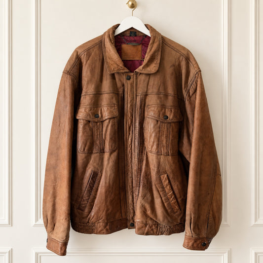 A brown leather jacket with a distressed finish, featuring a front zipper, two large chest flap pockets, and two lower hand-warmer pockets, hanging on a white hanger against a white paneled wall.