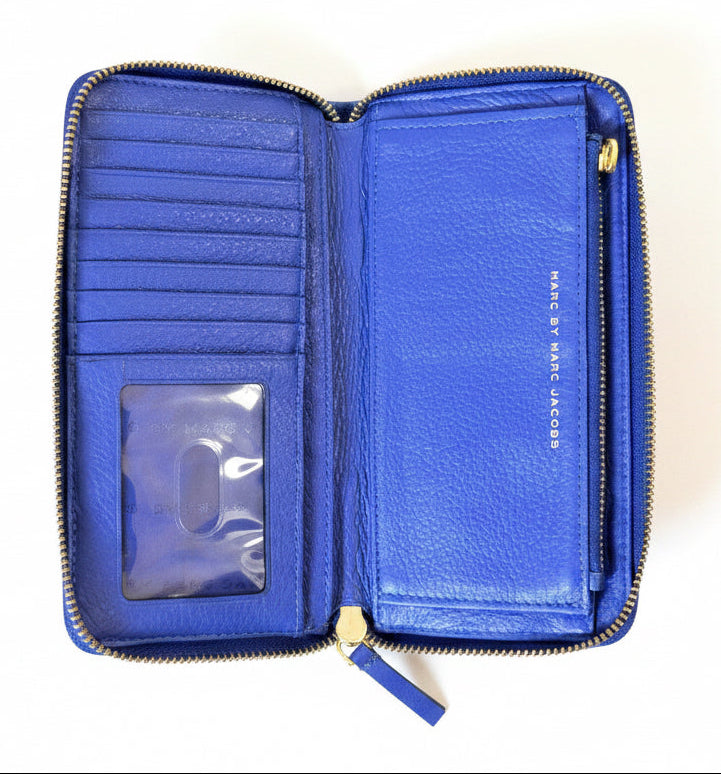 An open blue leather zip-around wallet with gold-tone zippers and a clear ID window, photographed from above on a green textured fabric background. The brand name 'MARC BY MARC JACOBS' is visible on the interior.