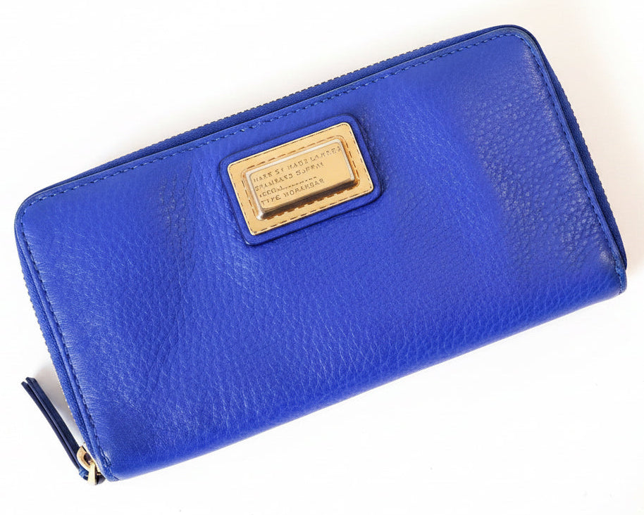 A bright blue pebbled leather zip-around wallet with a gold-tone metal nameplate, placed on a textured teal-green fabric background.