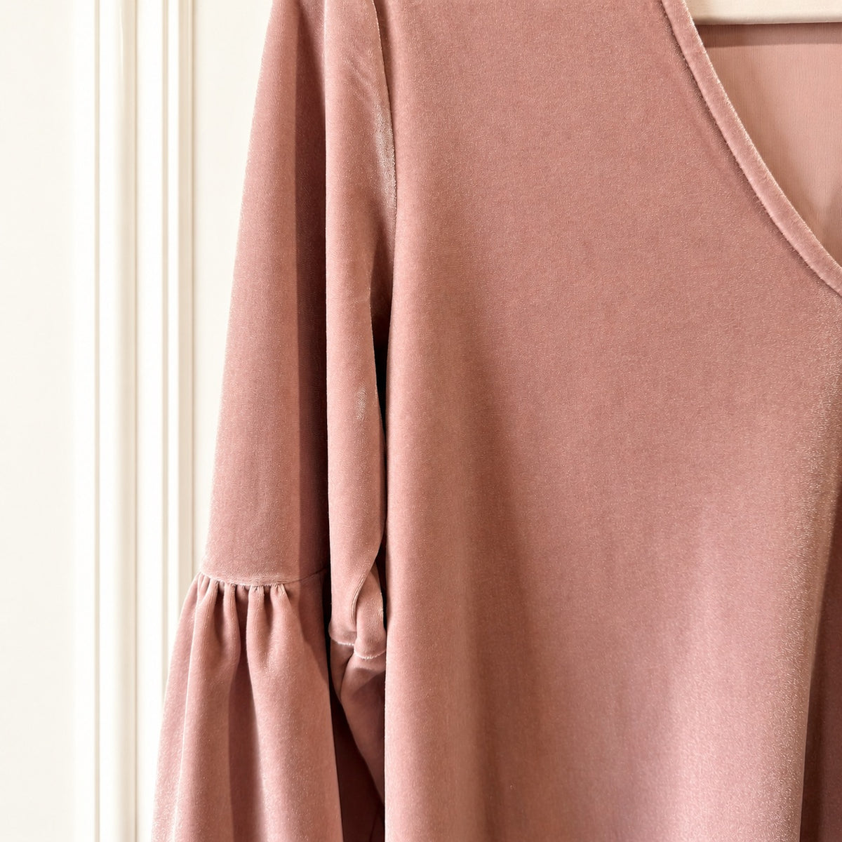A close-up of a dusty rose pink long-sleeve top with a V-neckline and gathered ruffle detail on the sleeve, hanging against a white paneled wall.