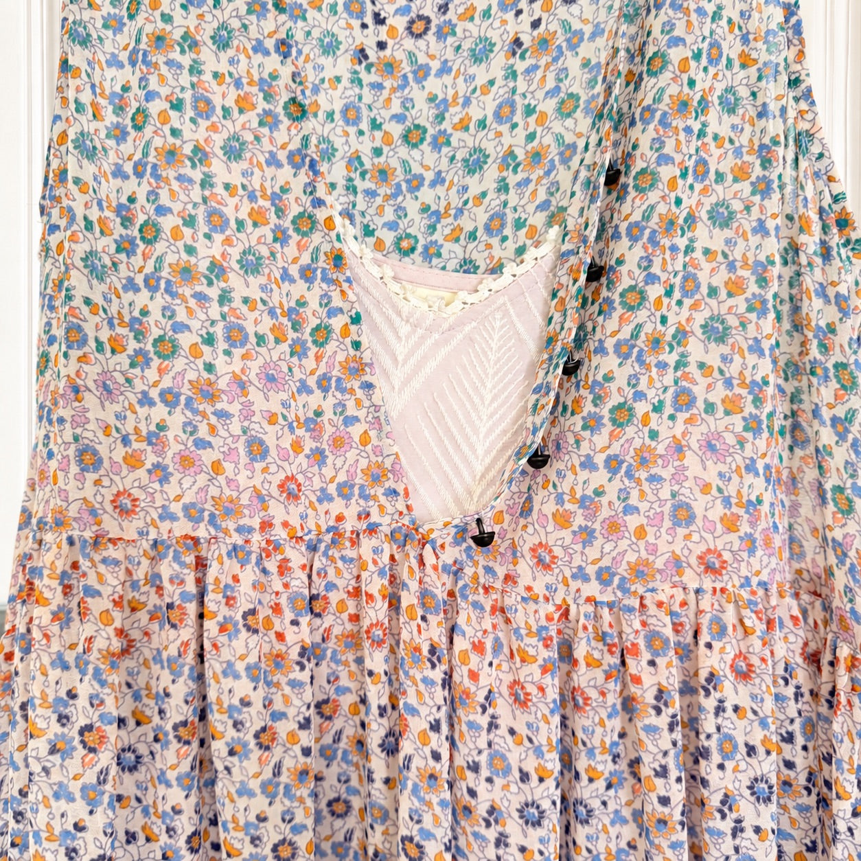 A close-up of a tiered bohemian-style dress featuring a dense multicolor floral print on a light cream base, with a scoop neckline and a lace-trimmed underlayer.