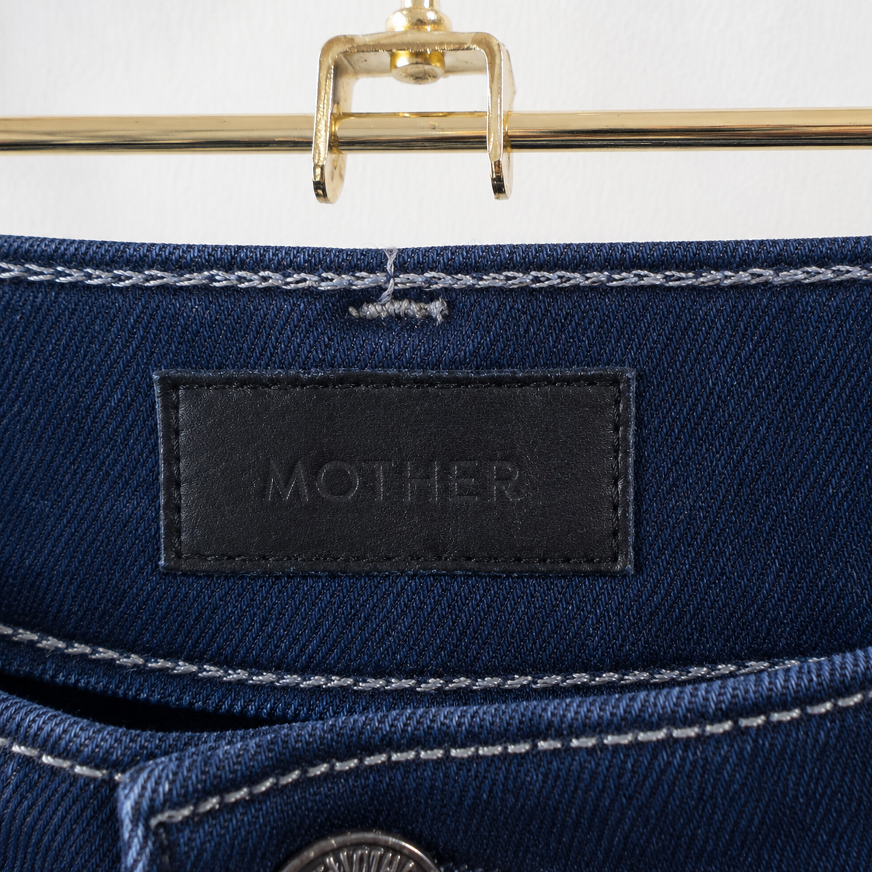 Close-up of the waistband of dark blue denim jeans featuring a black leather patch with the debossed brand name 'MOTHER'. The image shows white contrast stitching, a metal button, and a gold hanger hook at the top.