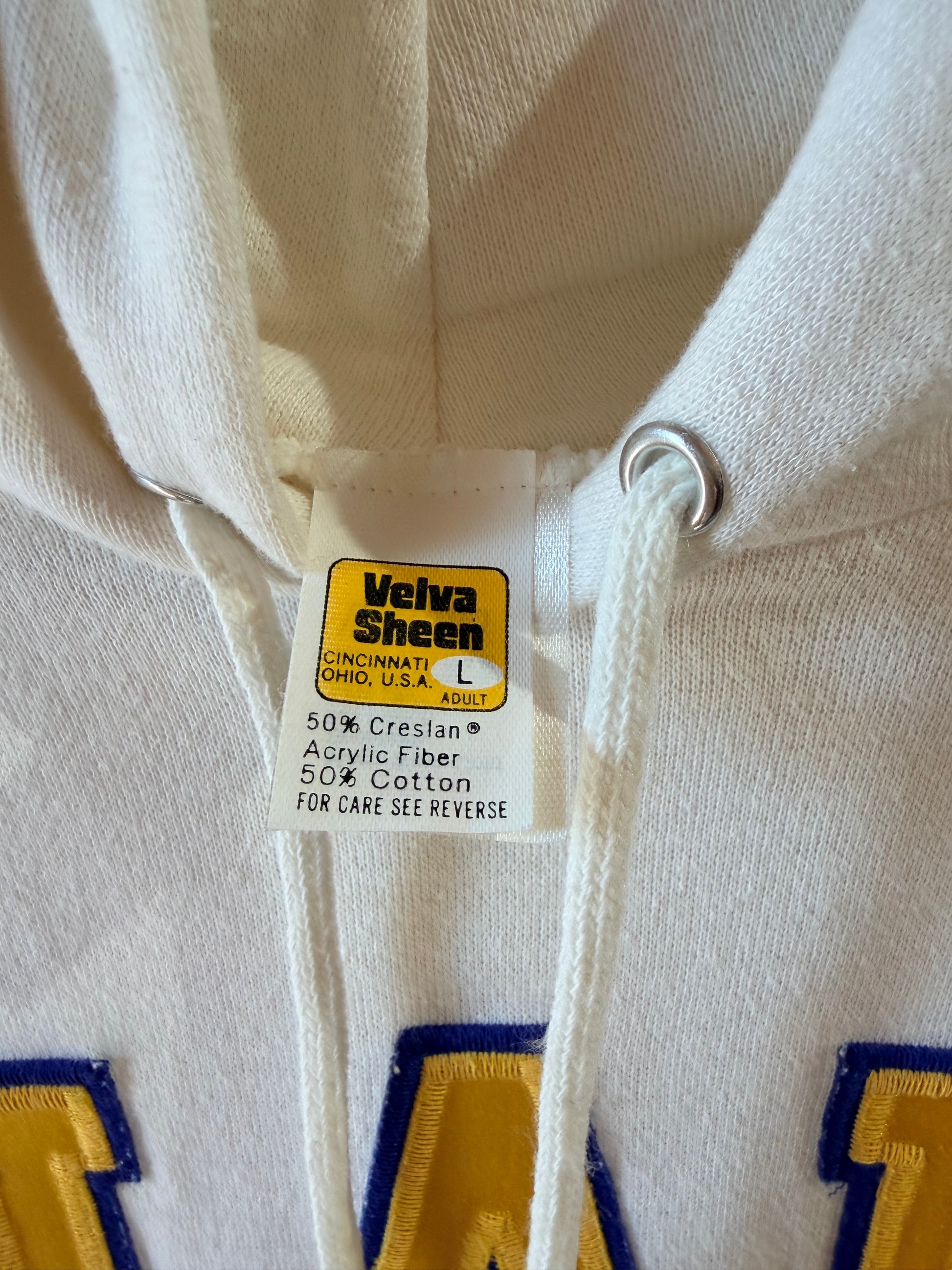 Close-up of a cream-colored hoodie showing the inner neck label which reads 'Velva Sheen' and '50% Crespian Acrylic Fiber 50% Cotton'. The yellow and blue lettering at the bottom is partially visible.
