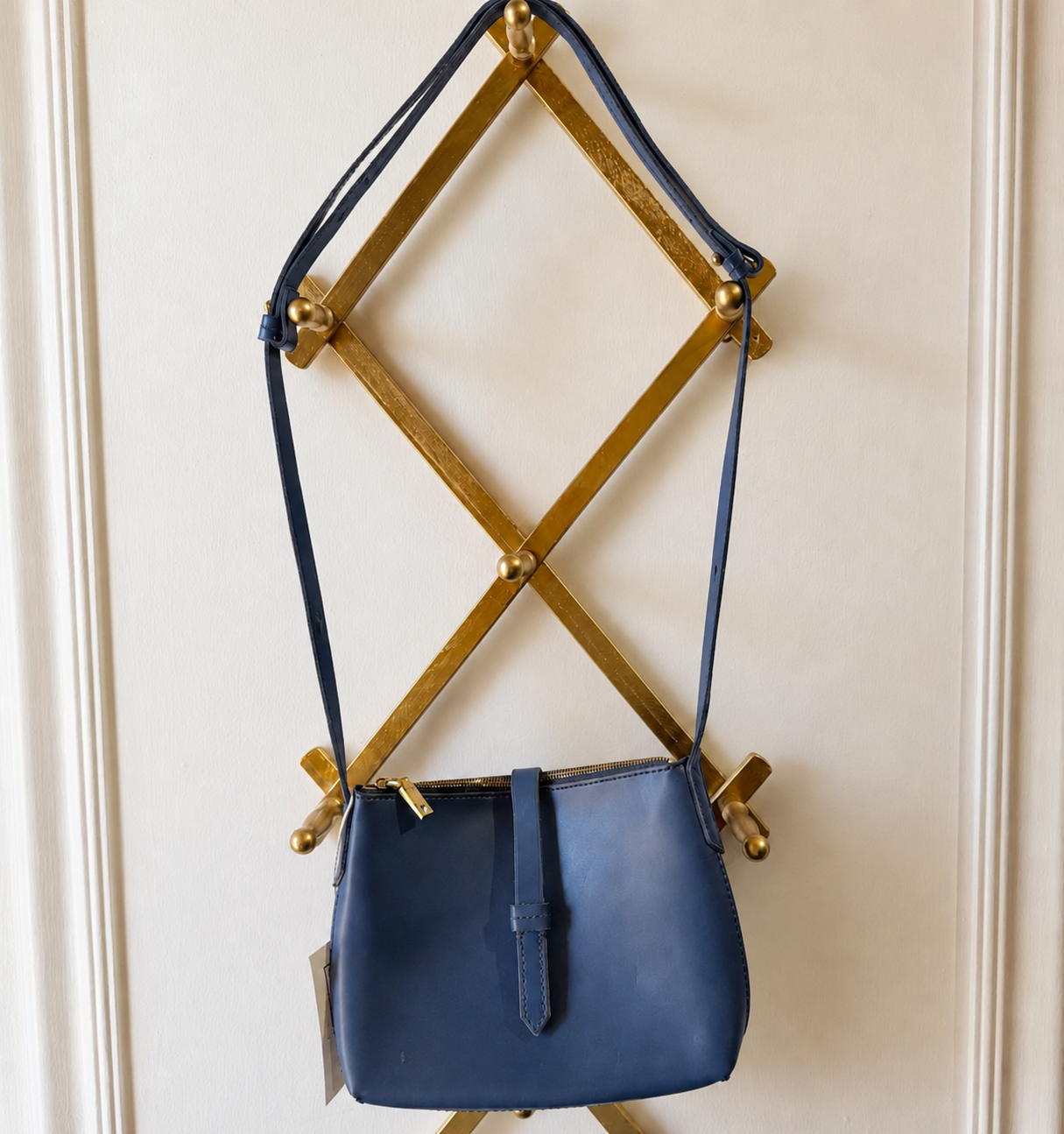 A navy blue leather shoulder bag with gold hardware, hanging from a gold geometric wall rack against a cream paneled wall.