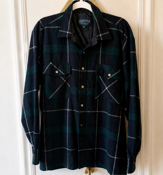 A vintage long-sleeve button-up shirt by Porter, featuring a dark navy blue and green plaid pattern with white lines, hanging on a wooden hanger against a white paneled door.