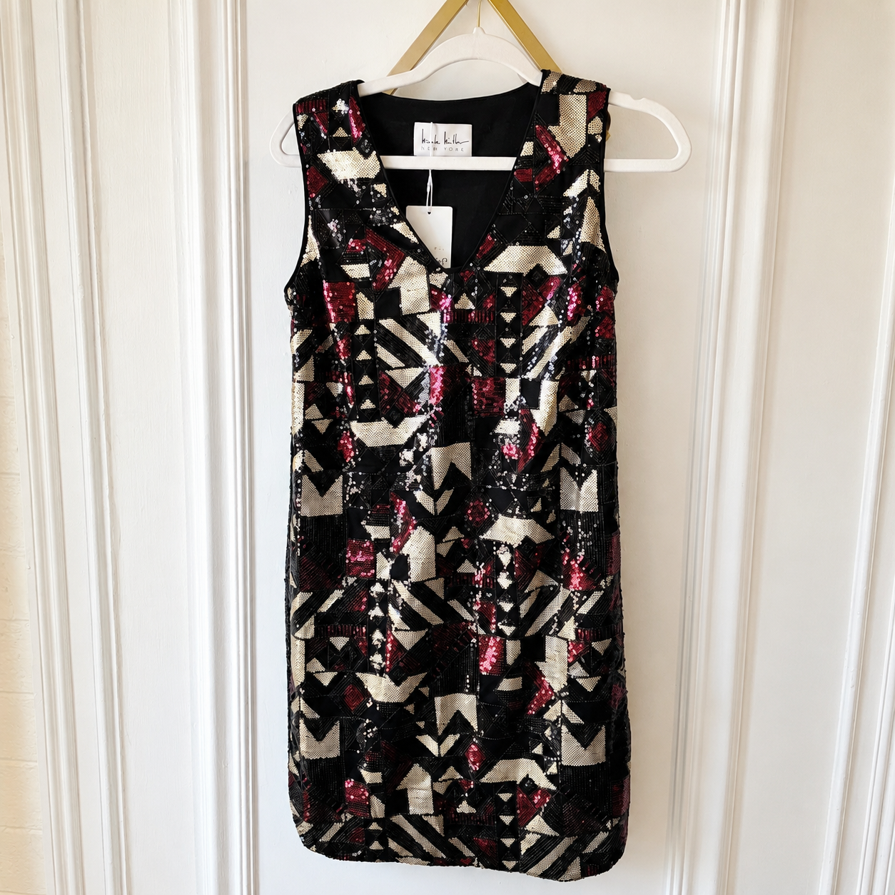 A sleeveless V-neck sequin dress hanging on a white hanger against a white paneled door. The dress features a geometric pattern of black, beige, and red sequins.