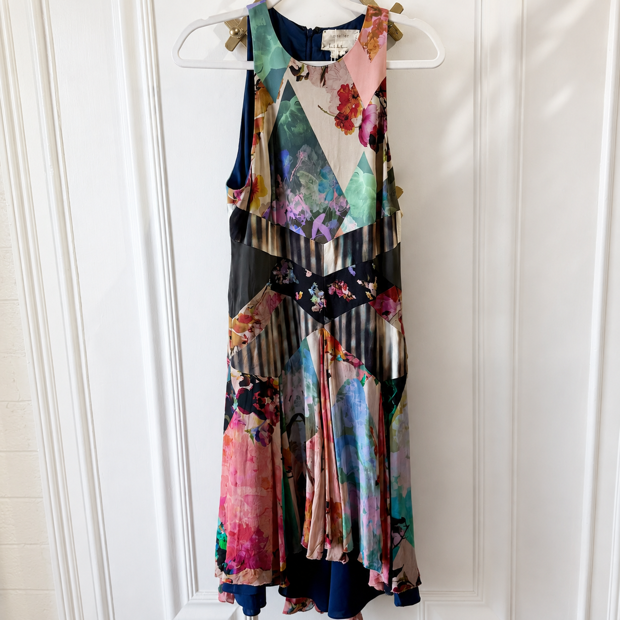 A sleeveless, multicolor patchwork-patterned dress with floral and geometric prints, hanging on a white plastic hanger against a white paneled door.