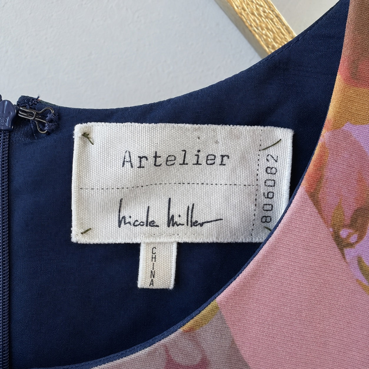 Close-up of a garment's inner neck label reading 'Artelier Nicole Miller' and 'CHINA', showing navy blue fabric, a zipper, and part of a pink floral patterned lining.