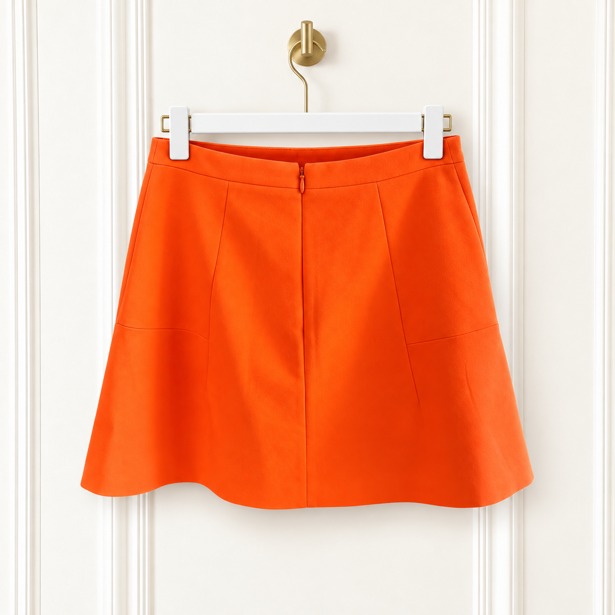 Bright orange A-line mini skirt with a zip closure and side pockets, hanging on a white hanger against a white paneled door.