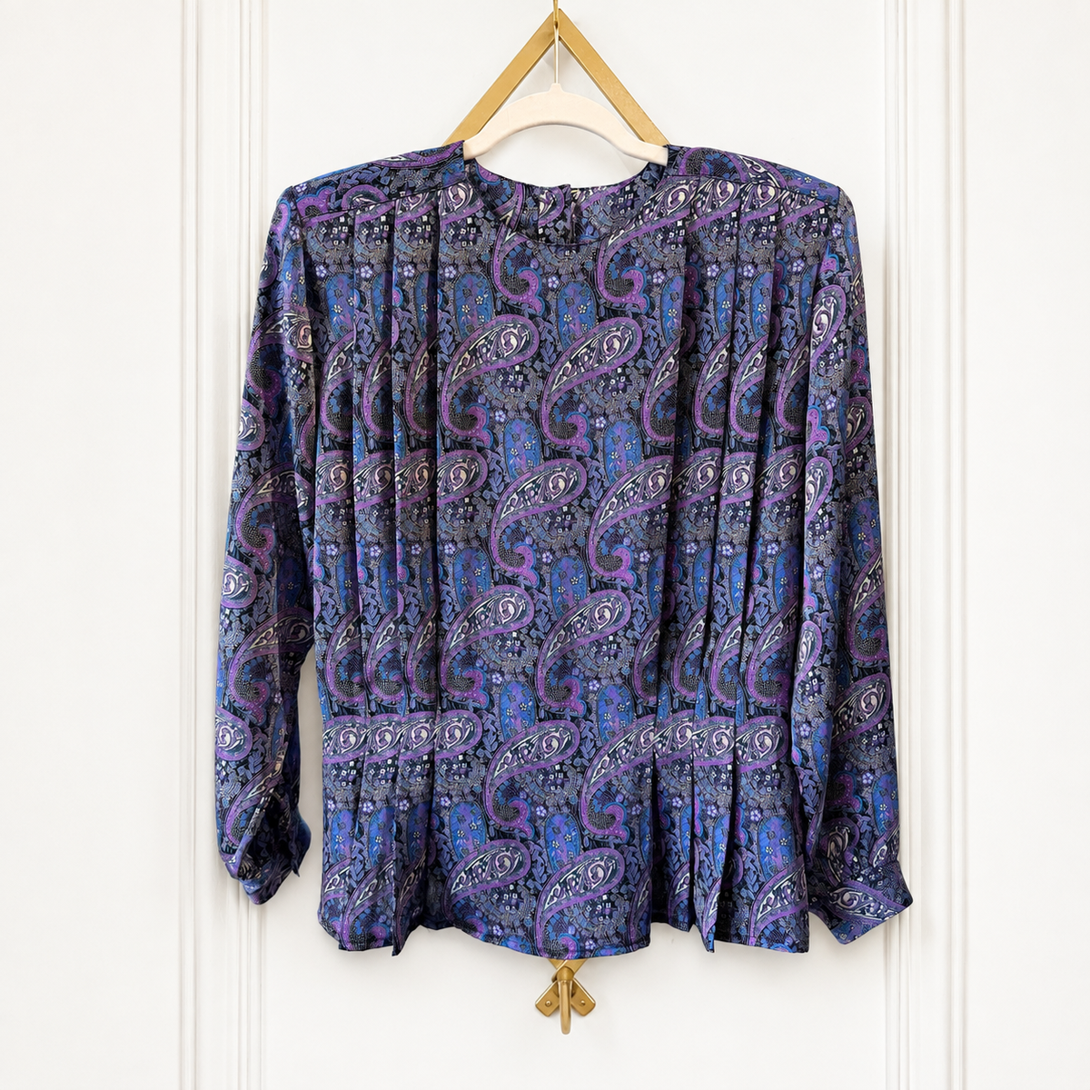 A long-sleeve blouse with a purple and blue paisley pattern, hanging on a wooden hanger against a white paneled wall.