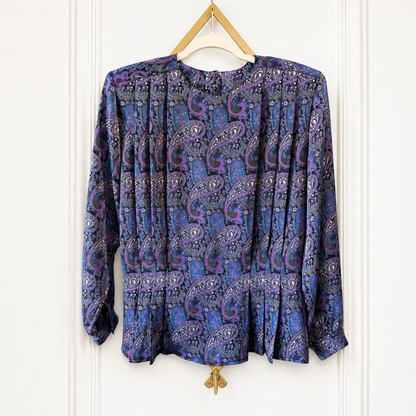 A long-sleeve blouse with a purple and blue paisley pattern, hanging on a wooden hanger against a white paneled wall.